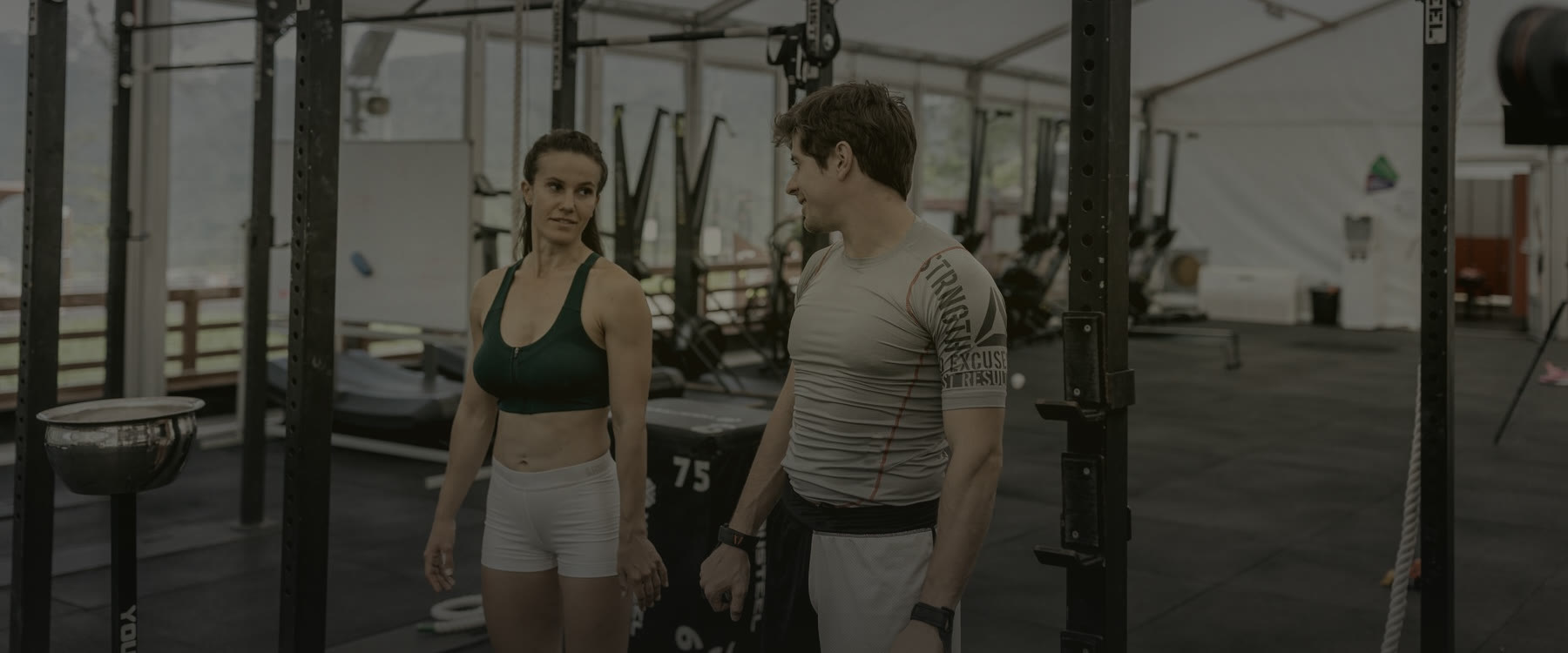 Deux coachs sportifs, une femme et un homme, se parlent au milieu d'un plateau de musculation, entre les cages à squat et les anneaux.